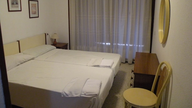 Hotellihuone, jossa on suuri sänky, yöpöytä lampun kanssa, tuoli ja ikkuna, jossa on verhot.