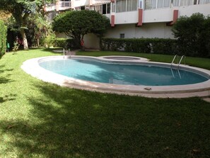 Una piscina amb una petita zona de solàrium, envoltada de gespa ben cuidada i alguns arbres.