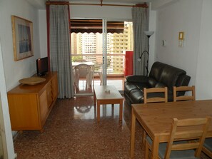 Ein Wohnzimmer mit einer Couch, einem Esstisch, einem Fernseher und einem Balkon mit Vorhängen.