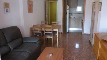 Sala de estar com sofá preto, mesa de jantar de madeira com cadeiras, cozinha ao fundo e porta que dá para outro quarto.