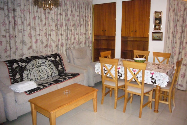 Una sala de estar con un sofá, una mesa de comedor de madera y sillas. Hay una ventana con cortinas, un reloj en la pared y una manta estampada con motivos florales sobre el sofá.