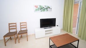 A living room with a flat-screen TV on a white cabinet, two wooden chairs, a wooden coffee table, and a sliding glass door with green curtains. - Oce3 3A in Benidorm With 2 Bedrooms and 1 Bathrooms (Benidorm)