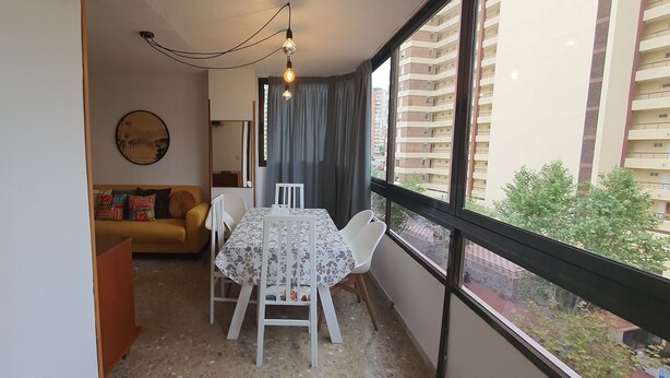 A small dining area with a table and chairs, a sofa, a wall-mounted painting, and a view of a residential building through large windows. - Mariscal 3 4J 2 Dormitorios (Benidorm)