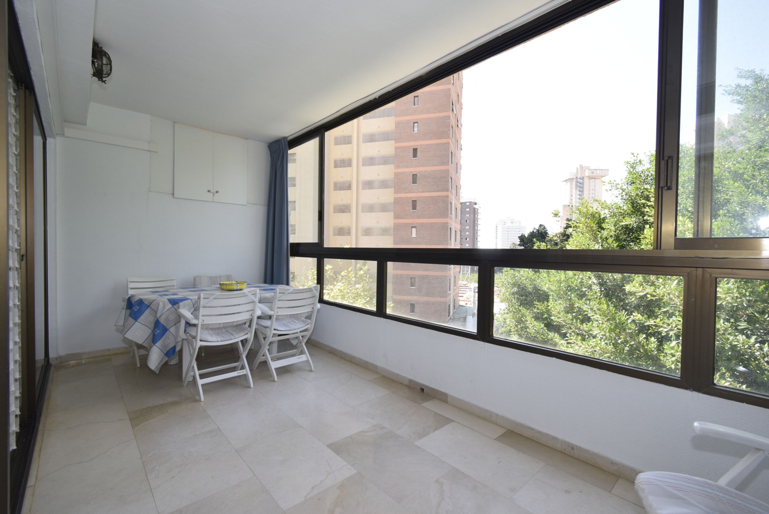 Un balcón con una mesa y sillas, vista de edificios y una ventana con marco negro.