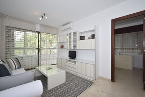 A modern living room with a white sofa, a glass coffee table, and a TV on a white cabinet. There is a large window with sheer curtains, and a door leading to another room.