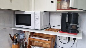 Eine kleine Küche mit weißen Schränken, einer Mikrowelle, einer Kaffeemaschine und einer Holzbrotkiste.