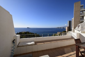 A balcony with a view of the sea, a white wall, and a table with chairs.