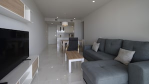 Una sala de estar moderna con televisor de pantalla plana, sofá gris, mesa de centro de madera y una zona de comedor al fondo.