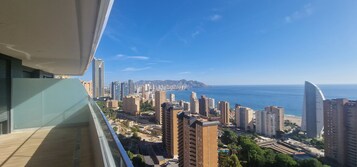 A high-rise building with a view of a coastal cityscape, including a beach and a mountain range in the distance.