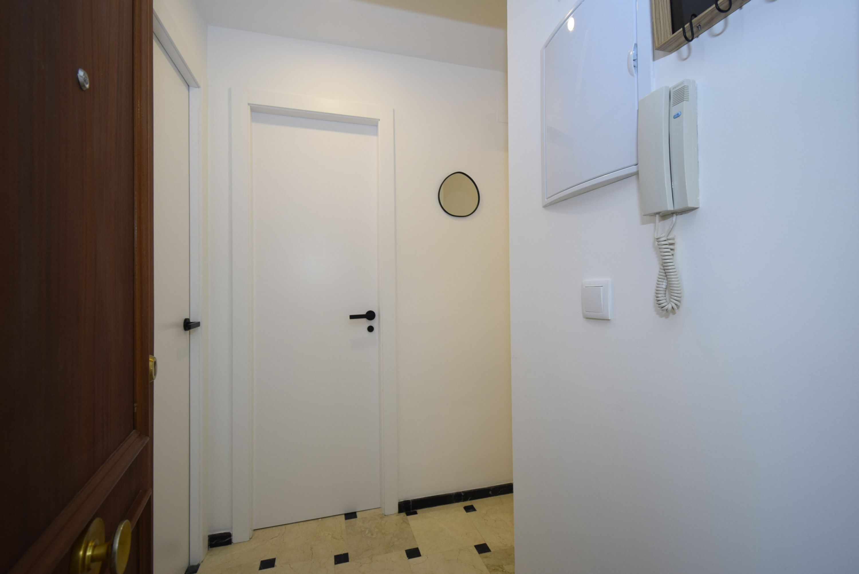 Un couloir avec une porte en bois, une porte blanche, un téléphone fixé au mur et une horloge murale.