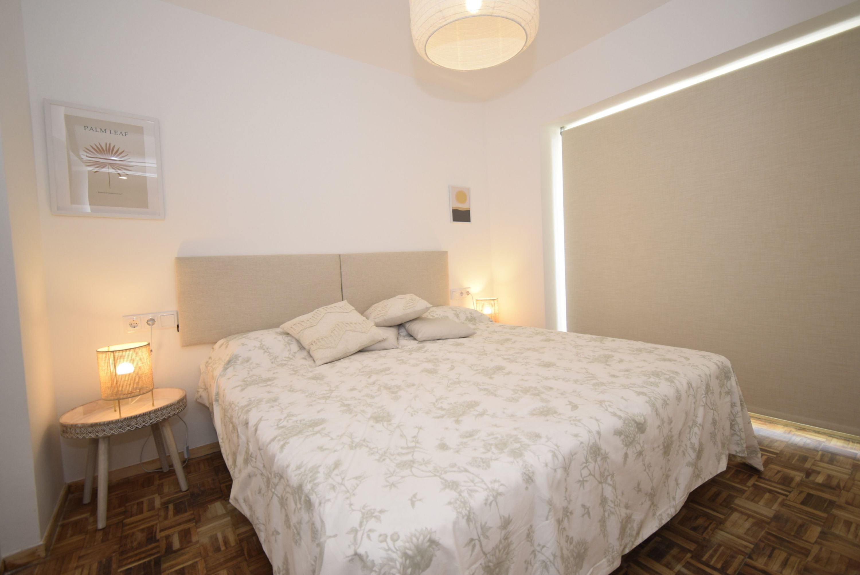 Un dormitorio con cama, una mesita de noche con lámpara, un cuadro en la pared y una ventana con persianas.