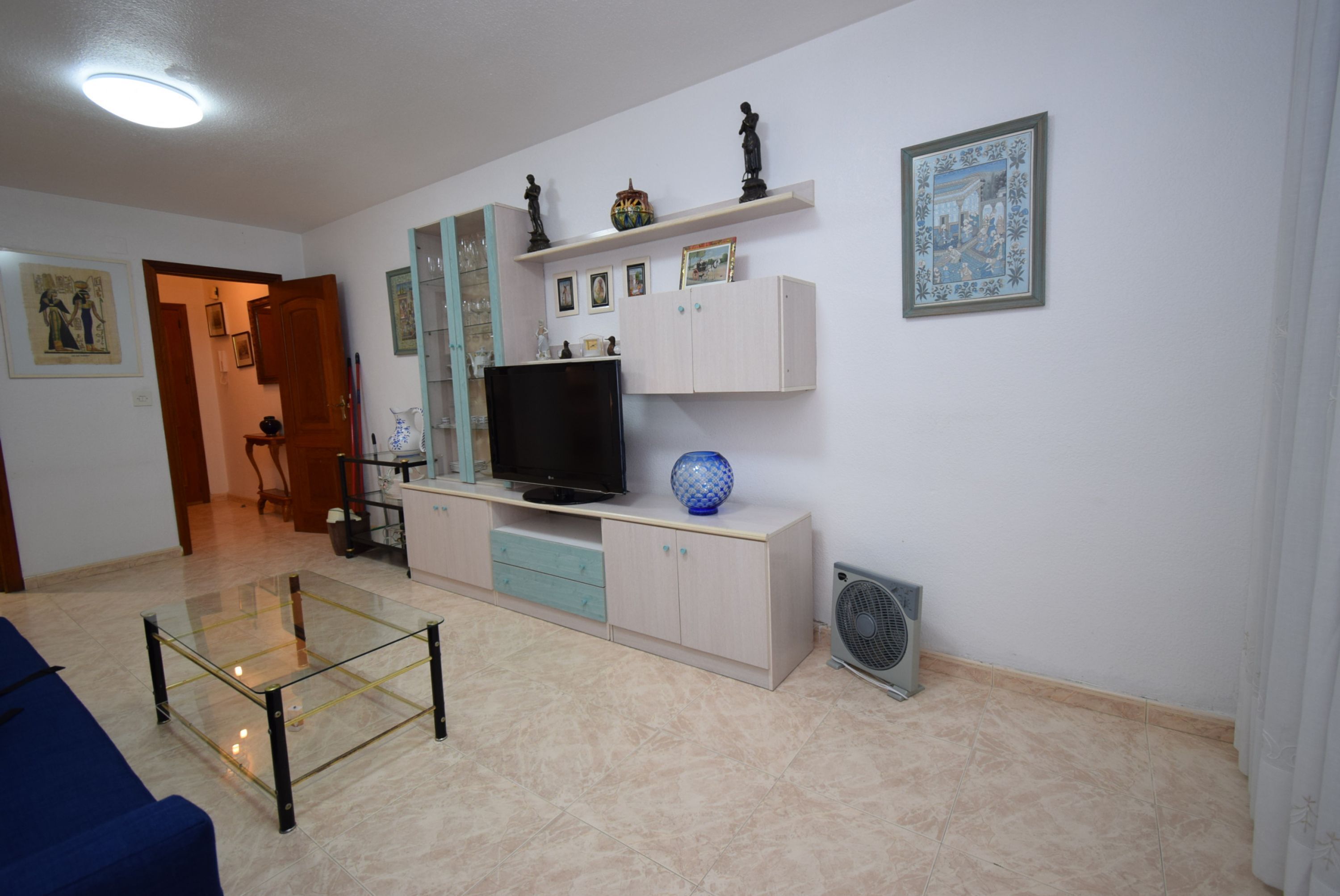 A living room with a glass coffee table, a television on a cabinet, a fan, and a blue vase.