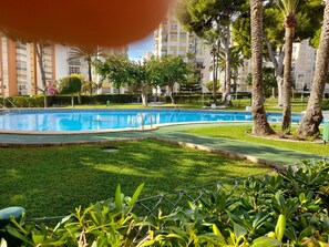 A swimming pool surrounded by well-maintained grass and trees, with a backdrop of residential buildings. - Urbanizacion Miramar 1057 (Benidorm)