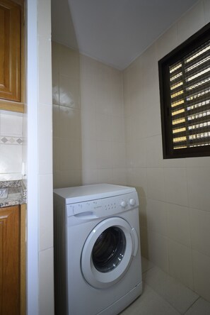 A compact laundry room with a front-loading washing machine, tiled walls, a wooden cabinet, and a window with blinds. - Castelmar 8B in Benidorm (Benidorm)