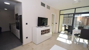 En moderne stue med et fladskærms-tv på et hvidt skab, et spiseområde med bord og stole, samt udsigt gennem store glasdøre til en udendørs balkon.