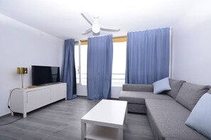 Uma sala de estar moderna com uma TV de tela plana, uma mesa de centro branca, um sofá cinza e um ventilador de teto.