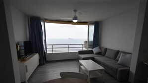 Un salon moderne avec un canapé, une table basse et un téléviseur posé sur un meuble. Une grande fenêtre offre une vue sur la mer.