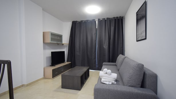 Un salon moderne avec un canapé gris, un téléviseur à écran plat posé sur un meuble en bois, une table basse et une grande fenêtre ornée de rideaux foncés.
