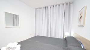 Une chambre d’hôtel avec un grand lit, une tête de lit, une fenêtre avec des rideaux, un miroir fixé au mur et un tableau encadré.