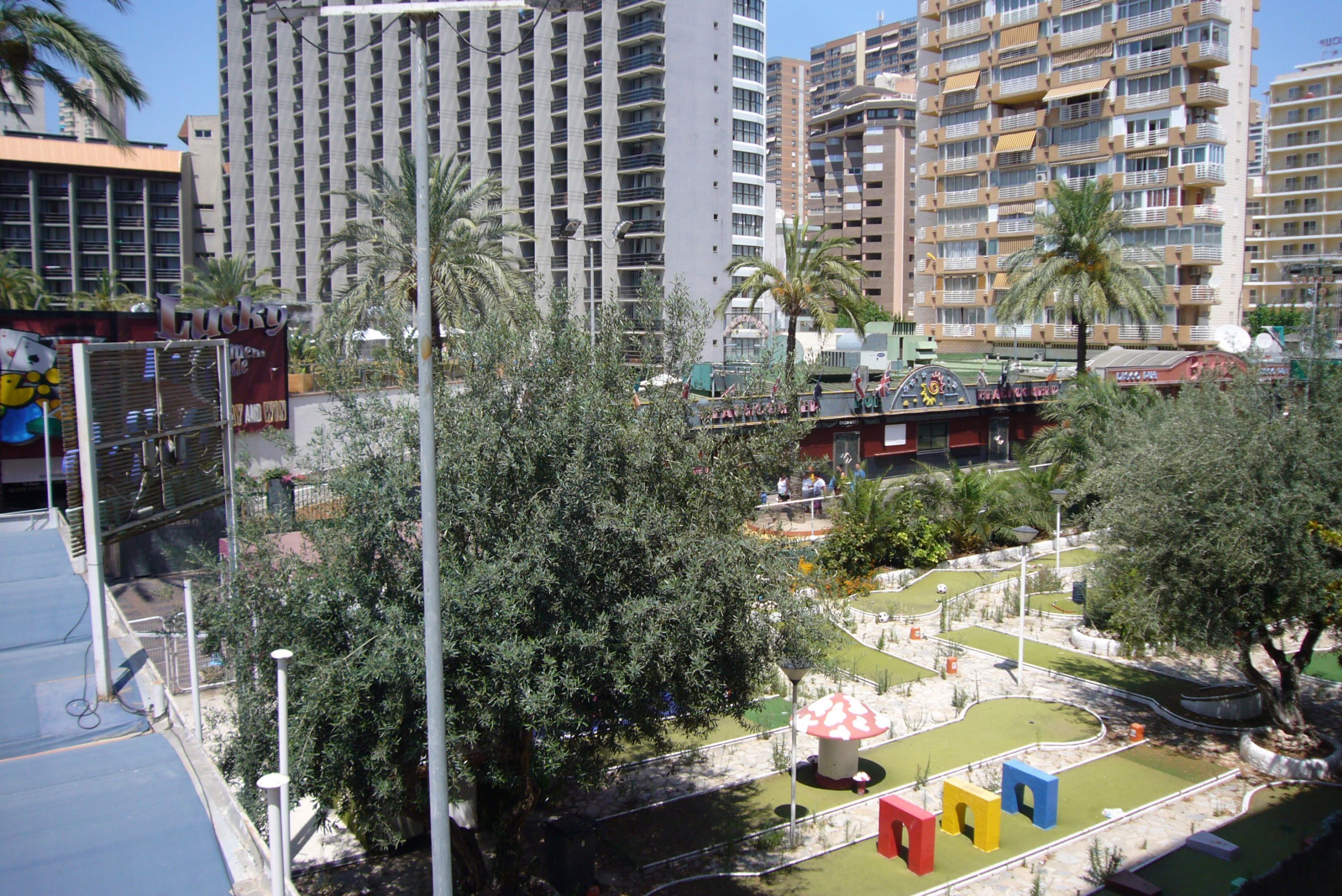 Un paysage urbain avec de hauts immeubles, un parc avec une petite aire de jeux et un grand bâtiment sur lequel on peut lire l’inscription « Lucky ».