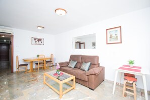 Ein Wohnzimmer mit braunem Sofa, einem Glastisch, einem Esstisch mit Stühlen, einem kleinen weißen Schreibtisch mit Stuhl und einem Spiegel an der Wand.