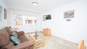 Ein Wohnzimmer mit einem braunen Sofa, einem Glastisch und einem Holzkabinett. An der Wand ist ein Fernseher angebracht und es hängt ein Kunstwerk mit Fahrradmotiv.
