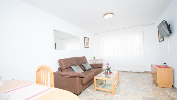 Ein Wohnzimmer mit braunem Sofa, einem Glastisch, einem Holznachttisch und einem an der Wand montierten Fernseher.