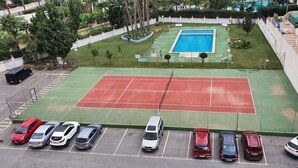 Ein Tennisplatz mit Pool, umgeben von einer Hecke und Bäumen. Autos sind in einer Reihe vor dem Platz geparkt.
