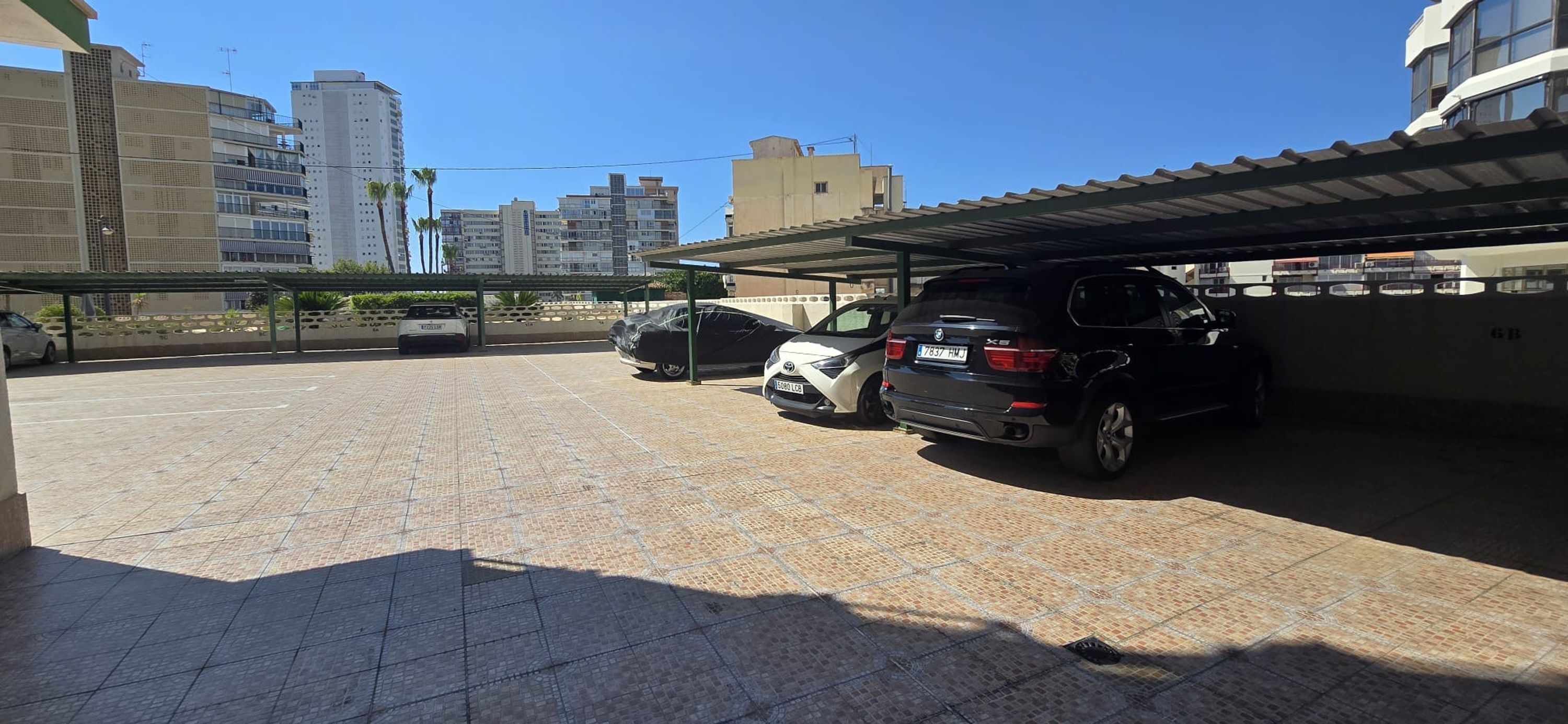 Un'area di parcheggio con diverse auto, una struttura coperta e edifici sullo sfondo.