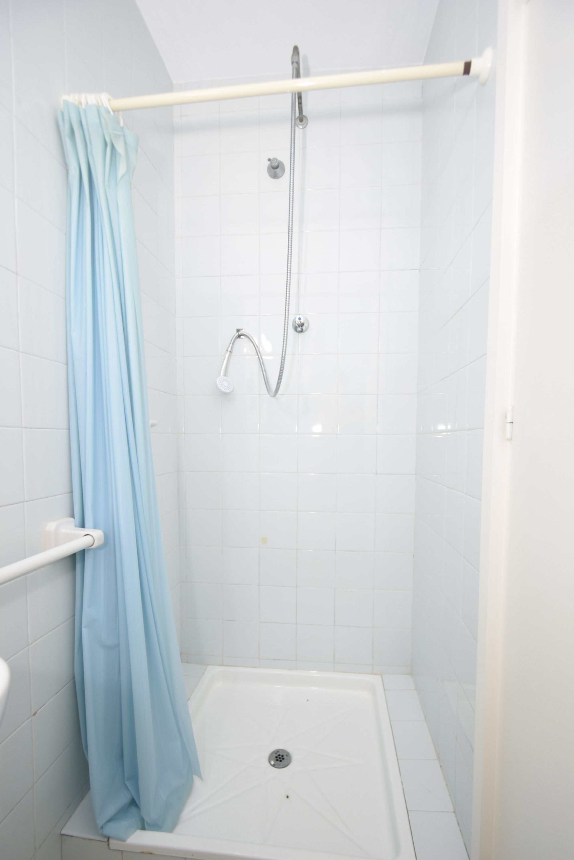 Un box de ducha pequeño con azulejos blancos, una cortina de ducha azul, un cabezal de ducha manual y un pasamanos blanco.