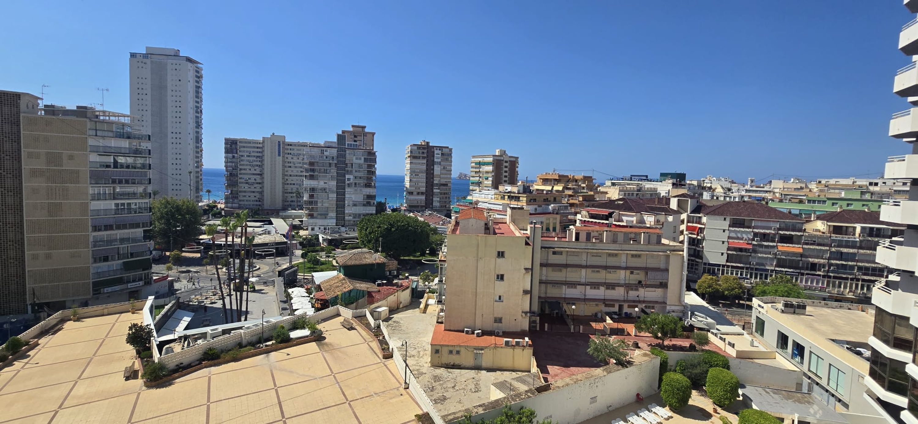 A cityscape with a mix of high-rise and low-rise buildings, a clear blue sky, and a view of the sea in the distance.