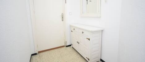 A small, white bathroom with a door, a vanity with drawers, a mirror, and a light switch on the wall.