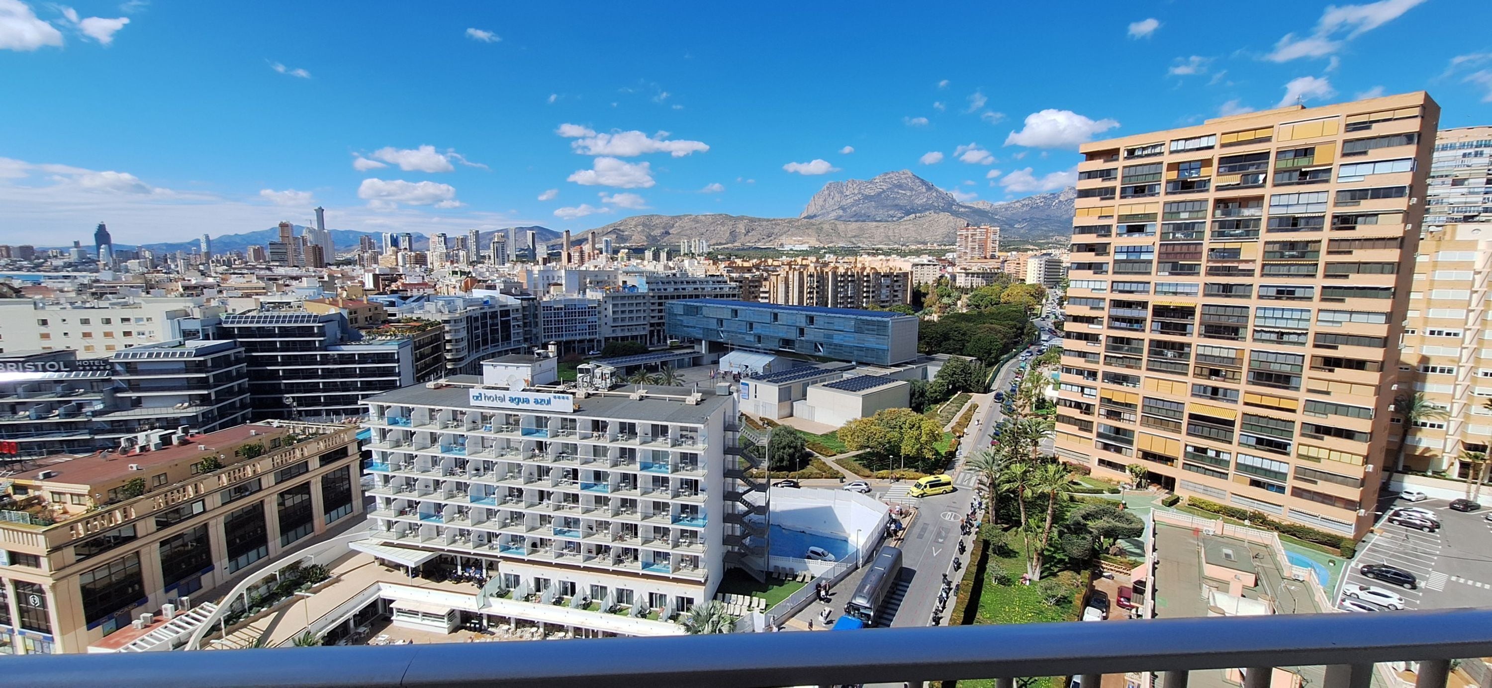 Un hôtel à étages, doté d’une piscine sur le toit et offrant une vue sur le paysage urbain et les montagnes en arrière-plan.