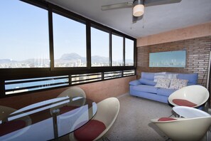 Ein modernes Wohnzimmer mit Blick auf die Stadt, einem Glastisch, zwei weißen Stühlen, einem blauen Sofa und einem Deckenventilator.