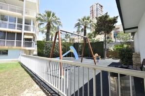 Ein Spielplatz mit Rutsche und Schaukeln, umgeben von einem weißen Zaun, Palmen und Wohnhäusern im Hintergrund.