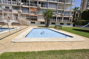 Ein Schwimmbecken mit einer gefliesten Terrasse, umgeben von einer Rasenfläche und einem mehrstöckigen Gebäude mit Balkonen.