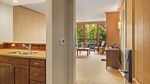 A kitchen with wooden cabinets and a granite countertop, leading to a living area with a large window and a view of greenery.
