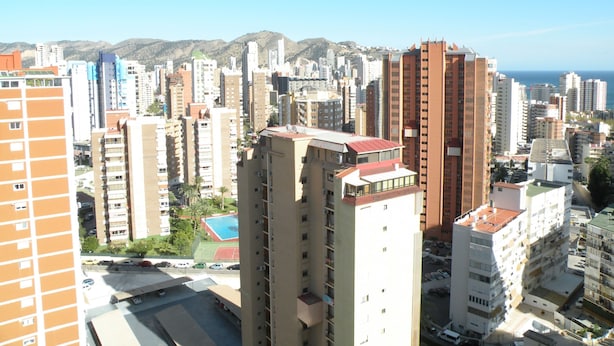 Un immeuble de grande hauteur, avec de nombreux étages, un espace de stationnement à l’avant et une vue sur le paysage urbain et les montagnes en arrière-plan.