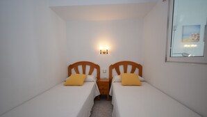 Een kamer met twee eenpersoonsbedden, elk met een houten hoofdbord en een geel kussen. Tussen de bedden staat een nachtkastje, en erboven hangt een wandlamp.