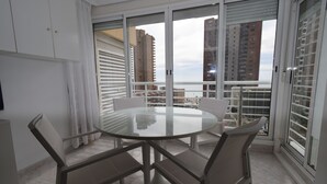Een moderne eetruimte met een glazen tafel, vier stoelen, grote ramen met zonwering en uitzicht op hoge gebouwen.