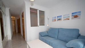Een woonkamer met een blauwe bank, een witte salontafel en een raam met jaloezieën. Er hangen vier ingelijste foto's aan de muur.
