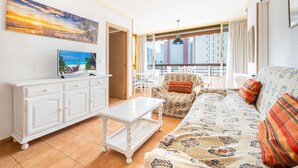 A living room with a sofa, a coffee table, and a TV on a white cabinet. There is a balcony with a view of buildings. - Acuarium 3 2F Estudio (Benidorm)