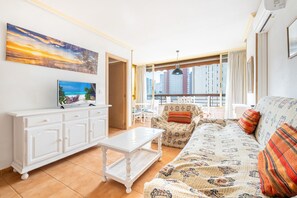 Ein Wohnzimmer mit einer Couch, einem Couchtisch und einem Fernseher auf einem weißen Schrank. Ein Balkon bietet Blick auf die Stadt.