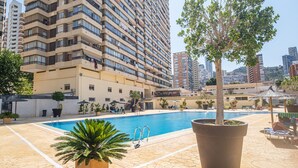 A swimming pool area with a large building in the background, surrounded by potted plants and lounge chairs. - Acuarium 3 2F Estudio (Benidorm)