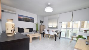 A modern living room with a sofa, coffee table, and dining area with chairs. There is a large window with blinds, a ceiling fan, and a vase on a table. - Marina Baixa 3D in Cala de Finestrat (Finestrat)
