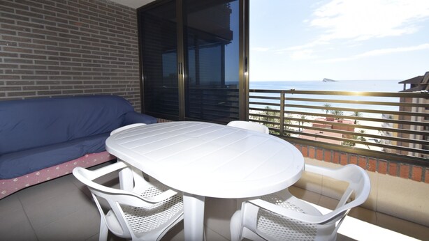 Een balkon met een witte tafel en stoelen, een blauwe bank en uitzicht op zee.