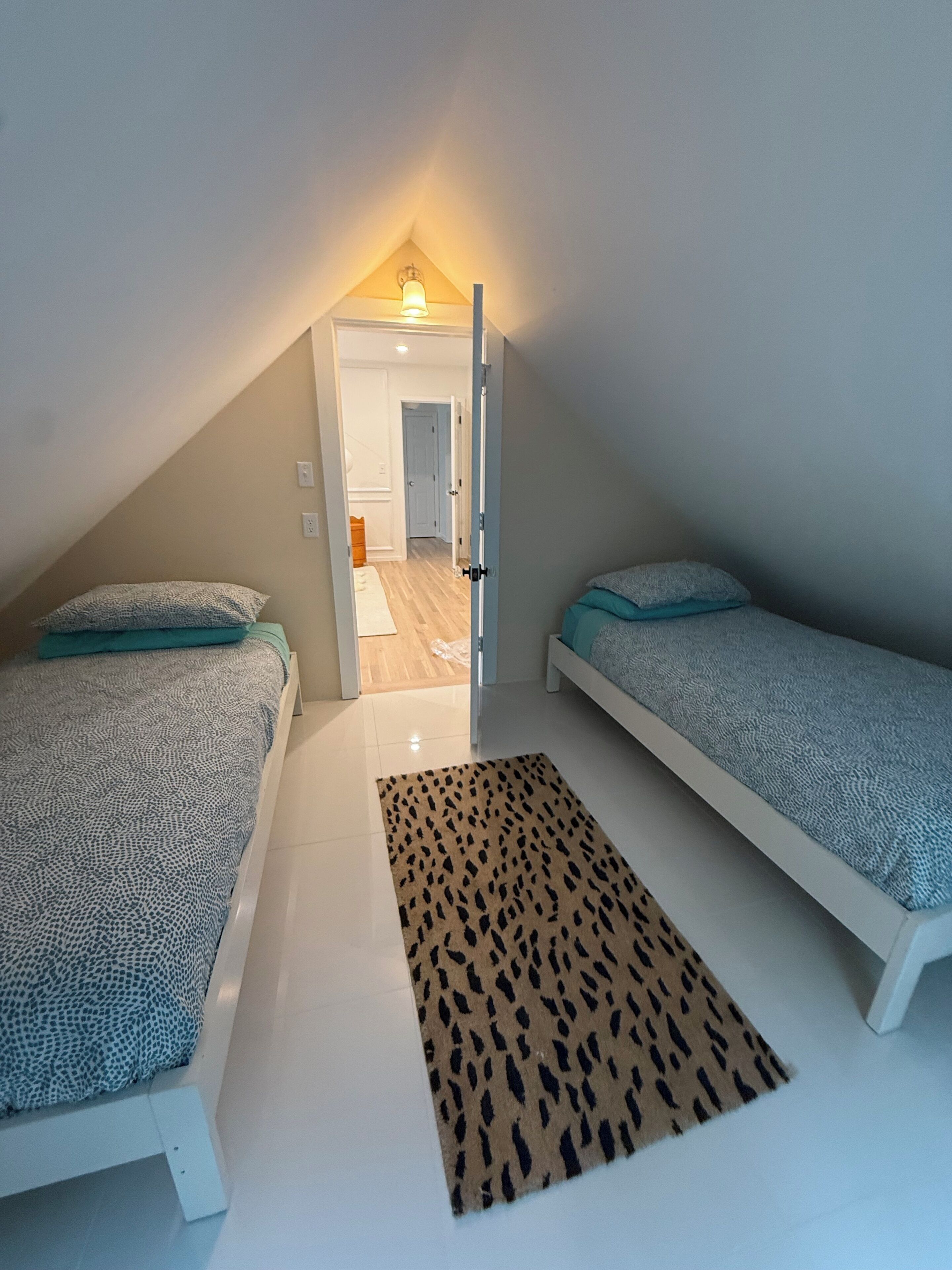 Une petite chambre mansardée de forme triangulaire, avec deux lits simples, un tapis à motifs et une porte donnant sur un couloir.
