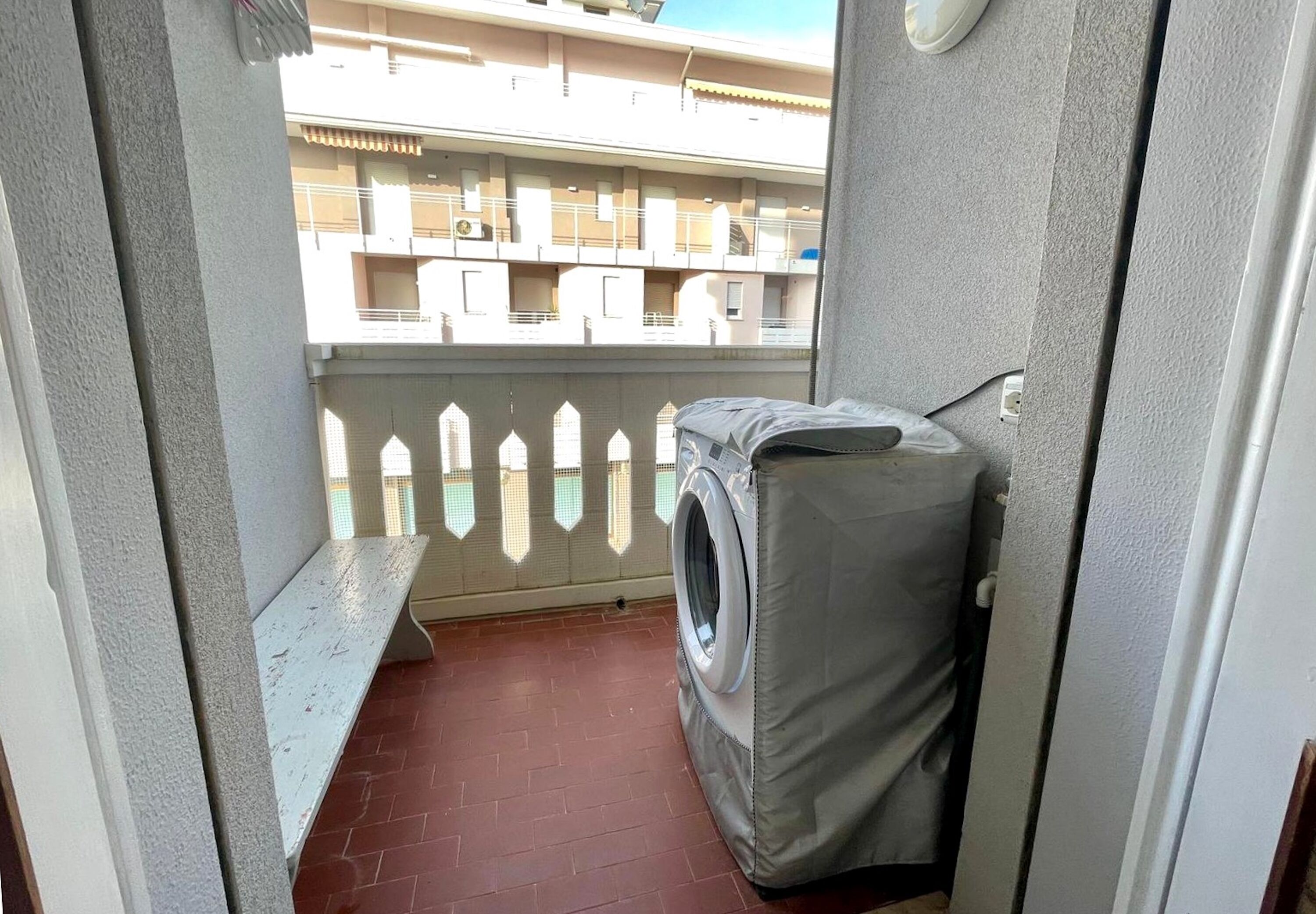 Un balcón con lavadora cubierta, una banca y vistas a un edificio con balcones.
