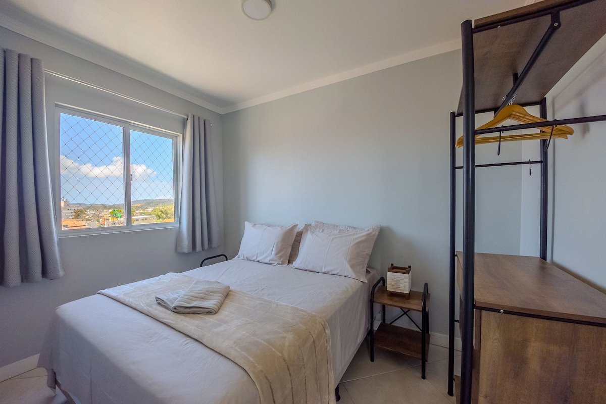 A hotel room with a bed, a small bedside table, a wooden shelf, and a window with curtains.