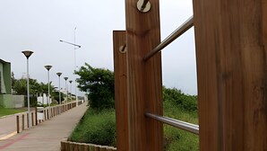 Un puente peatonal de madera con barandillas metálicas, que conduce a un camino con farolas y follaje.